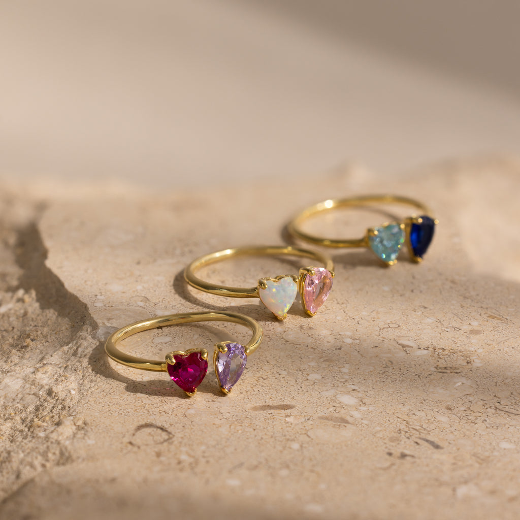 The Love Toi et Moi Ring in 18K Gold features four heart-shaped gemstones in different colors, displayed on a textured beige surface—ideal for stacking or wearing as a romantic statement piece.