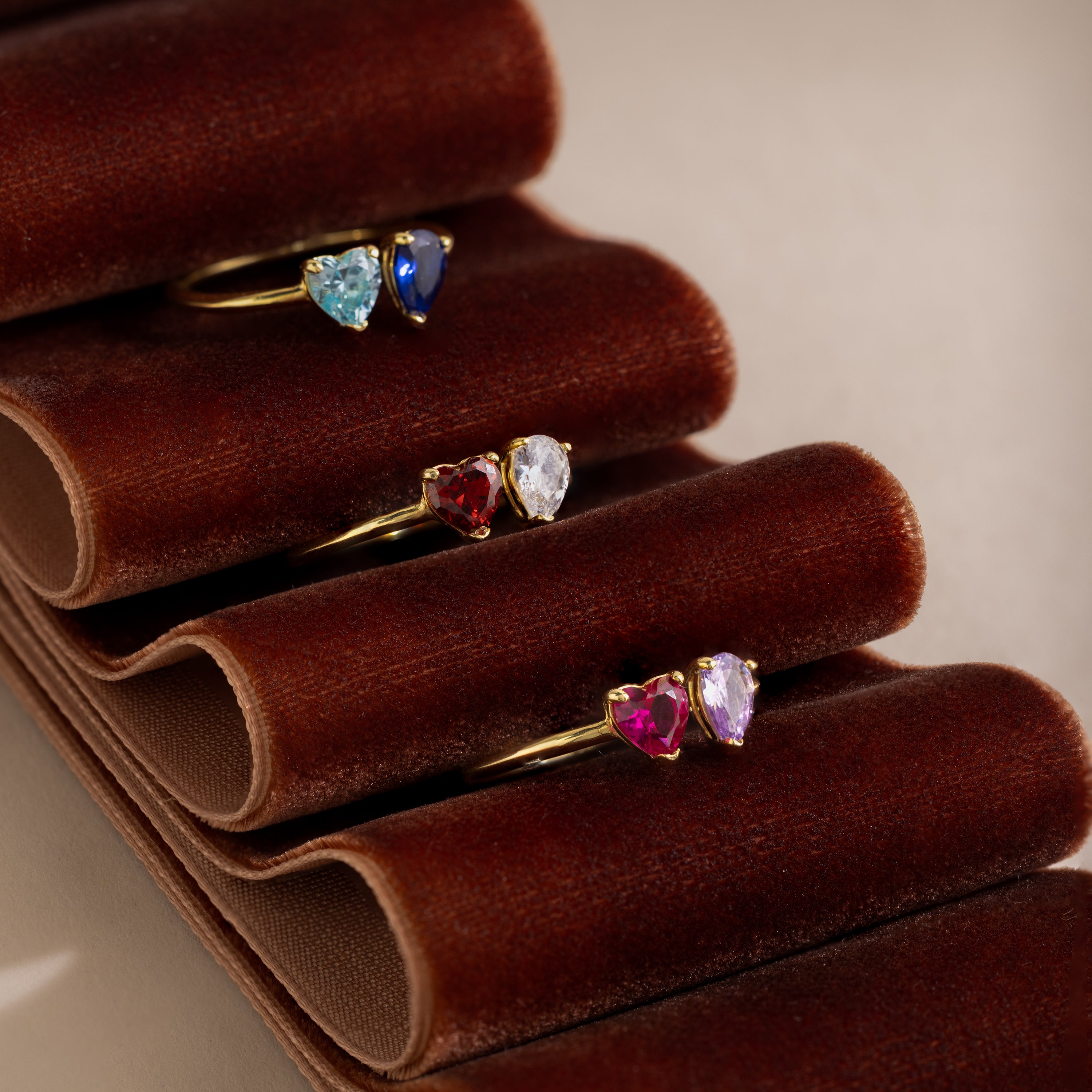 The Love Toi et Moi Ring features four gold stacking bands with colorful heart-shaped gemstones, displayed on a brown velvet ring holder.