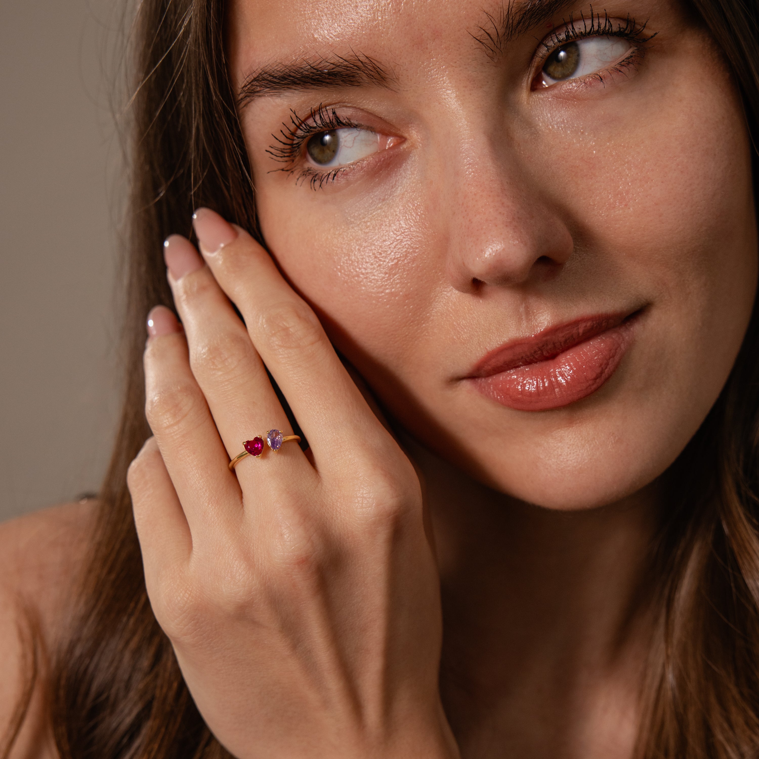 A woman with long brown hair touches her face, wearing the Love Toi et Moi Ring in 18K Gold with a striking red gemstone on her finger.