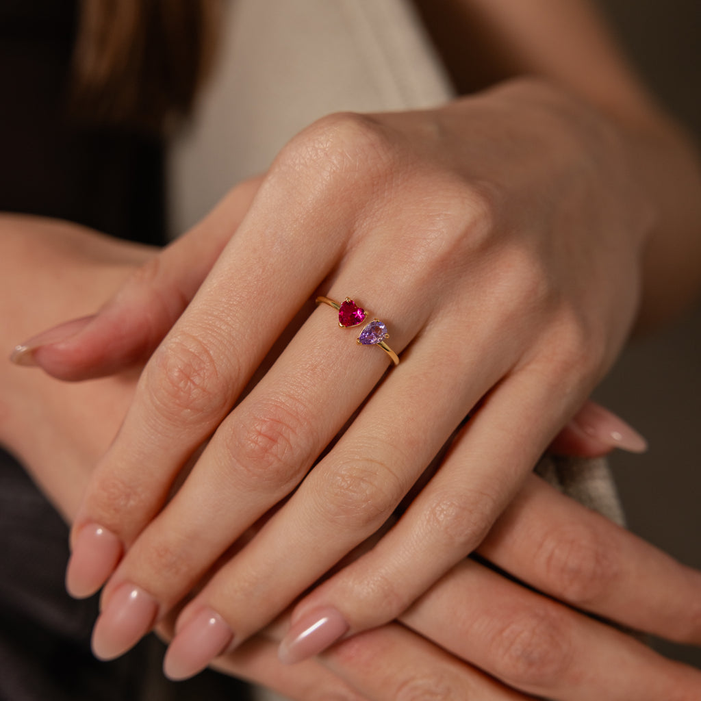 A hand with manicured nails wears the Love Toi et Moi Ring in 18K Gold, featuring a red and a purple gemstone.