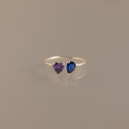 The Love Toi et Moi Ring in Silver features two heart-shaped gemstones—one purple and one blue—set side by side and shown against a plain neutral background.