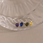 The Love Toi et Moi Ring in Silver features four heart-shaped gemstones in purple, blue, green, and yellow, elegantly stacked on a clear glass surface.