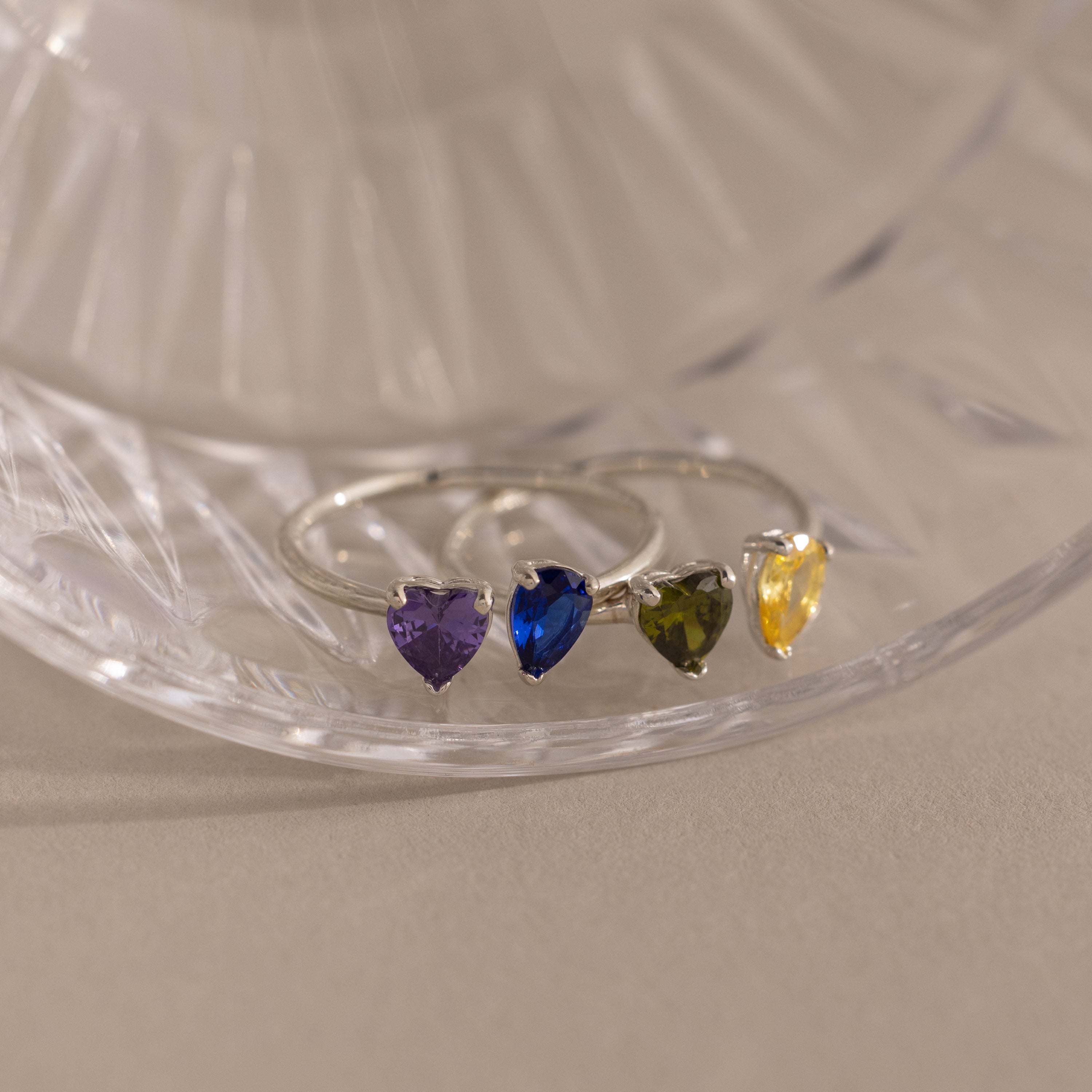 The Love Toi et Moi Ring in Silver features four heart-shaped gemstones in purple, blue, green, and yellow, elegantly stacked on a clear glass surface.
