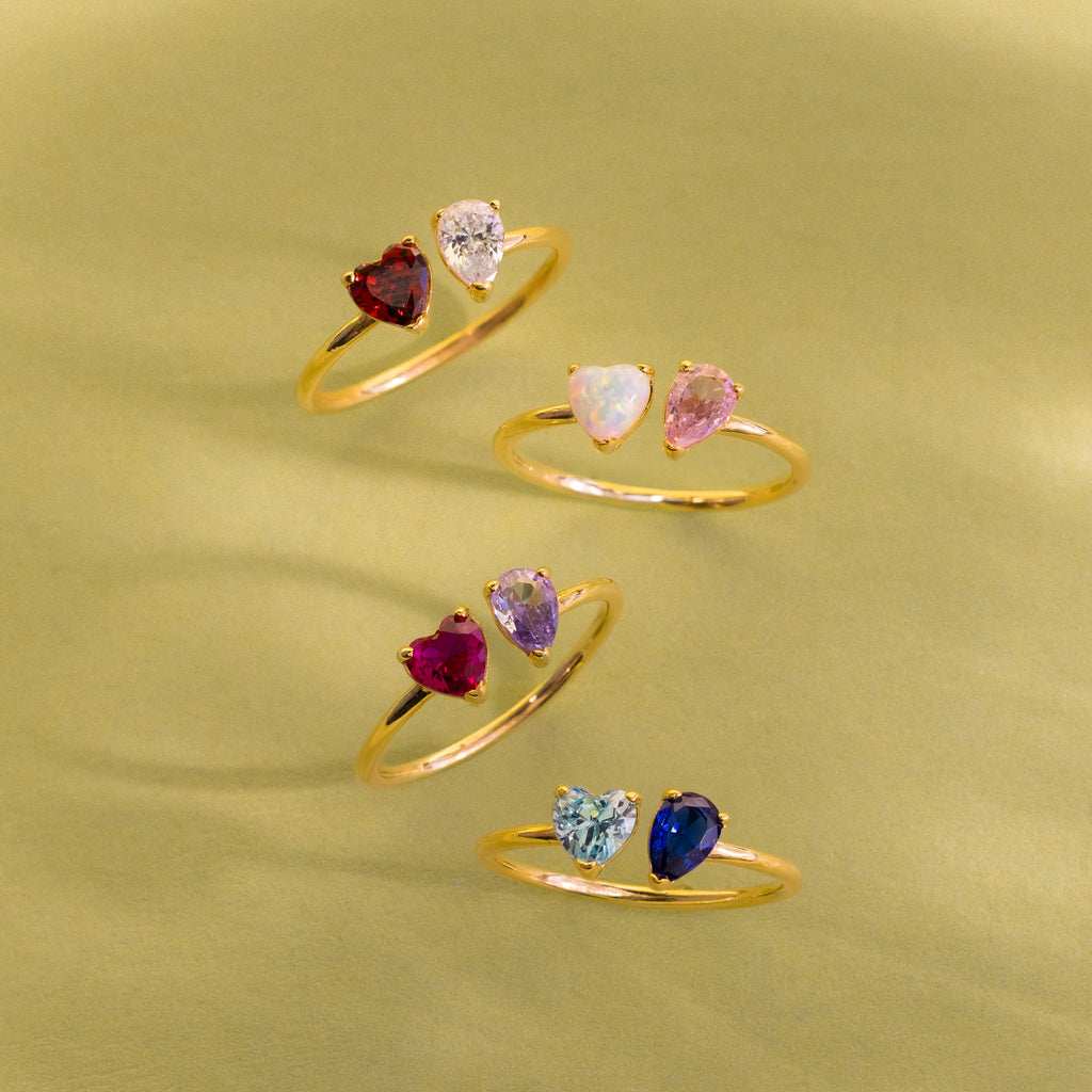 The Love Toi et Moi Ring in 18K Gold features four stacking rings with paired heart-shaped gemstones in varied colors, showcased against a soft yellow-green backdrop.