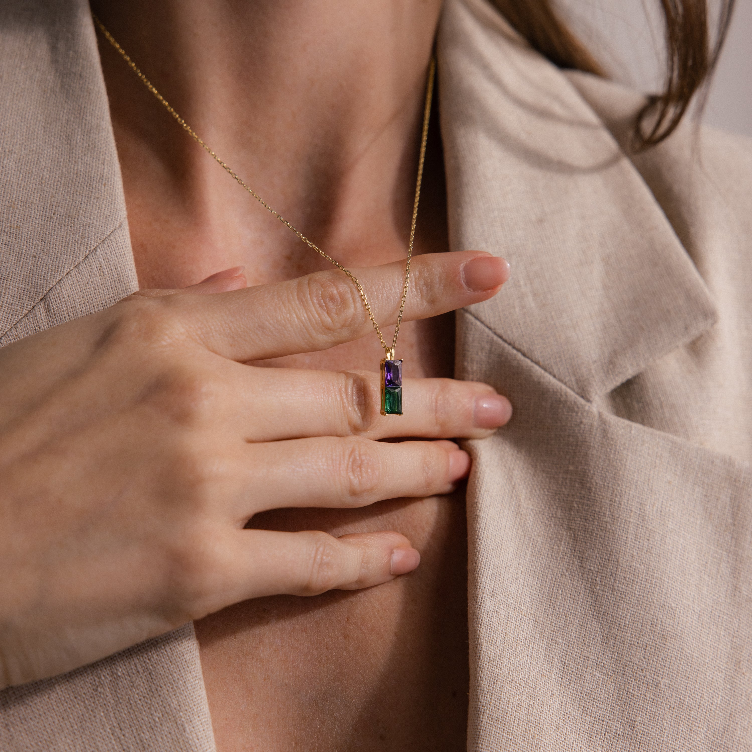 A woman in a beige blazer holds the Duo Birthstone Name Necklace in 18K Gold, featuring a rectangular purple and green gemstone pendant.