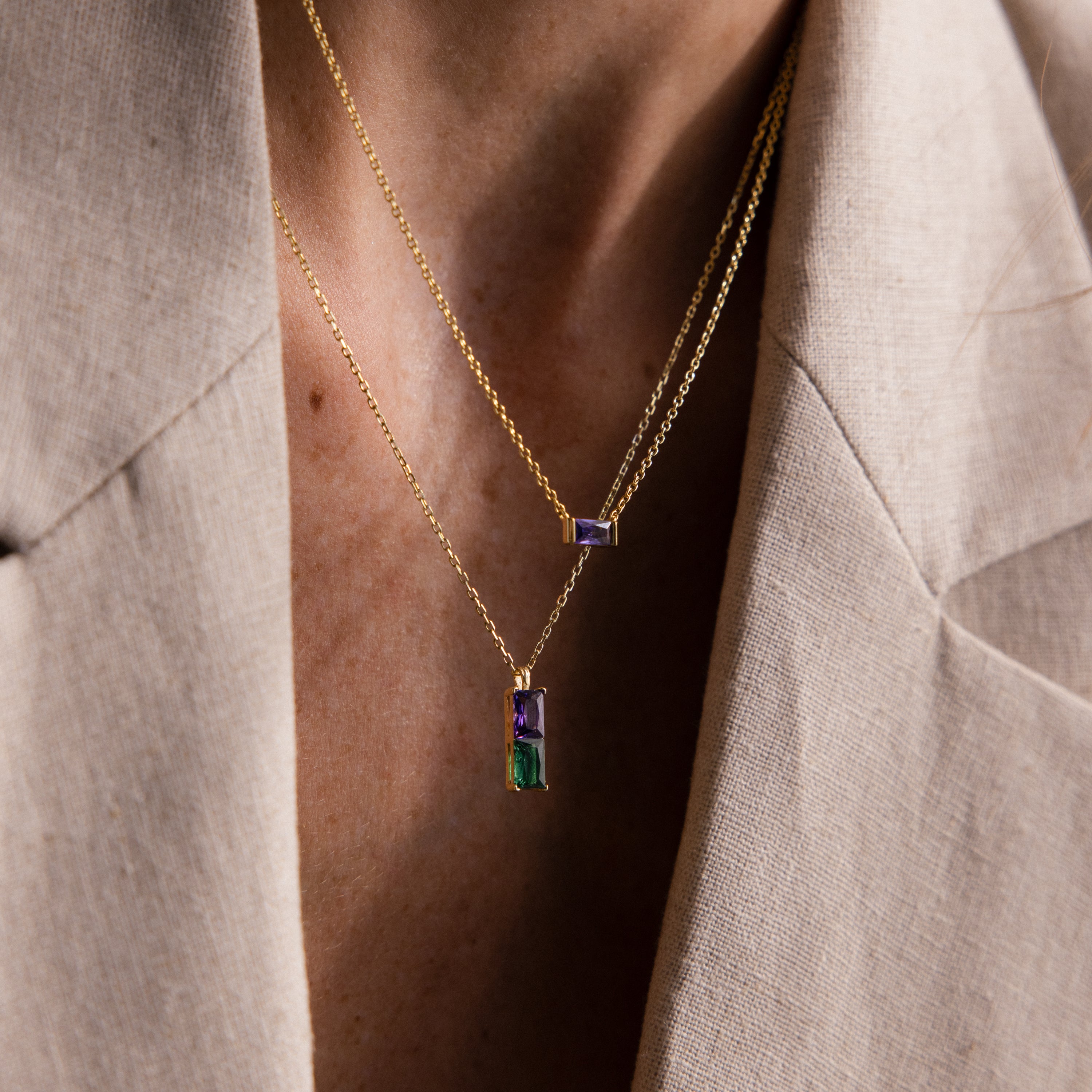 A person in a beige blazer wears layered gold necklaces, including the Duo Birthstone Name Necklace in Rose Gold featuring colorful rectangular stones and custom name engraving.