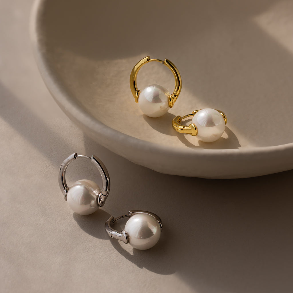 Two pairs of Bold Round Pearl Hoops earrings in gold and silver are displayed on a beige ceramic dish and surface.