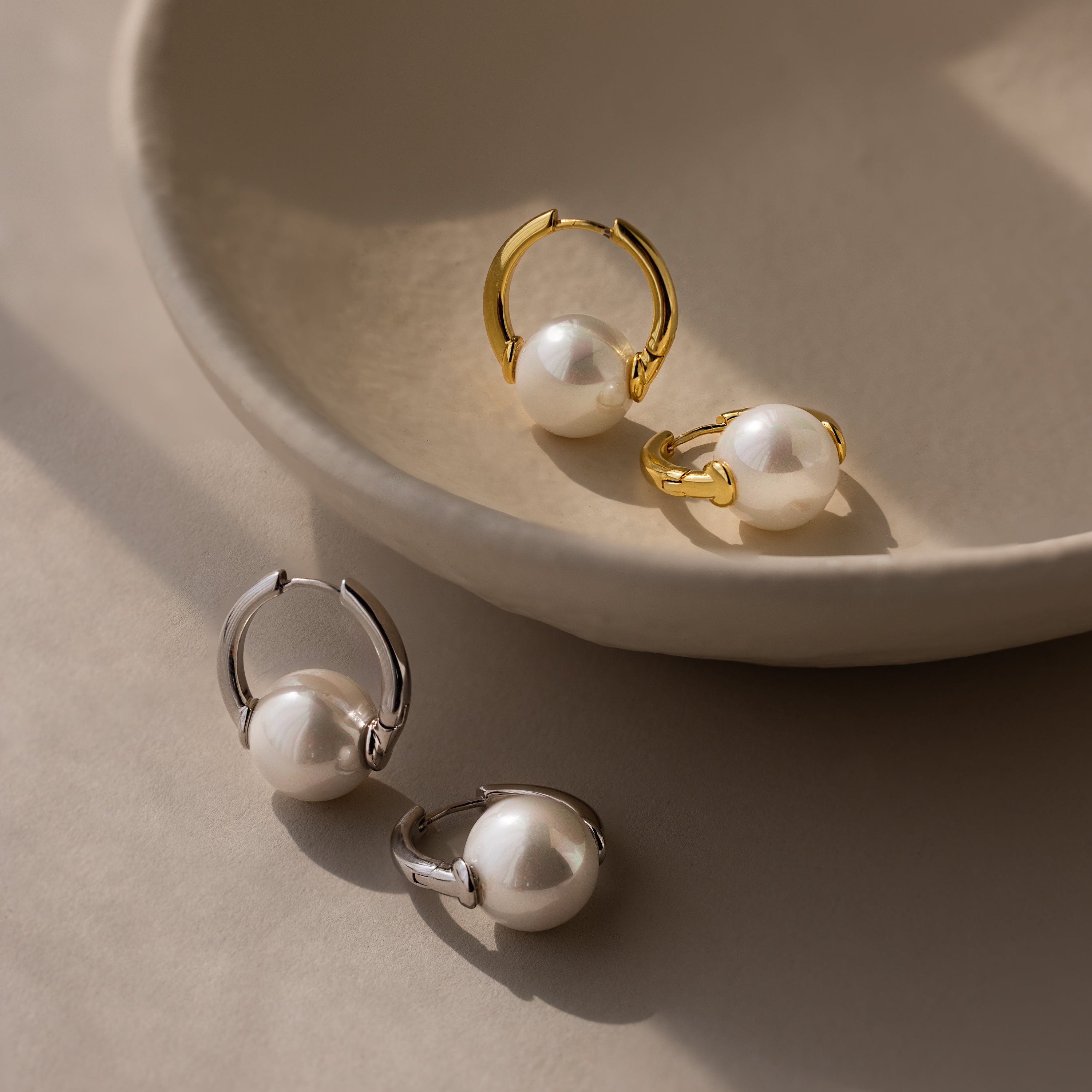 Two pairs of Bold Round Pearl Hoops earrings in gold and silver are displayed on a beige ceramic dish and surface.