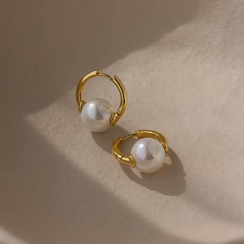 The Bold Round Pearl Hoops in 18K Gold feature large white pearls on gold hoops, displayed on a smooth beige surface—ideal statement jewelry for pearl lovers.