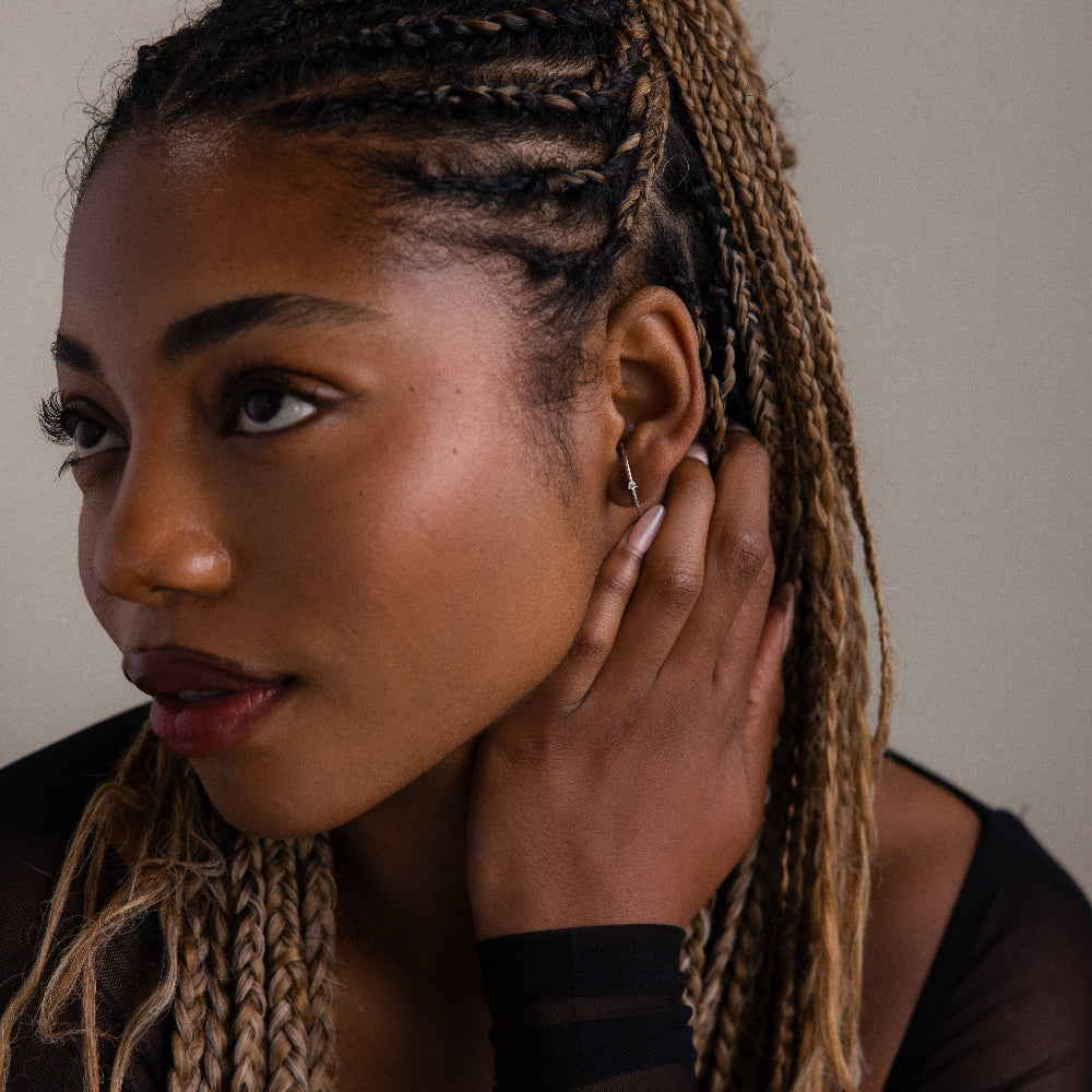 Woman with braided hair and a diamond studded ear jacket style earring touching her ear against a neutral background