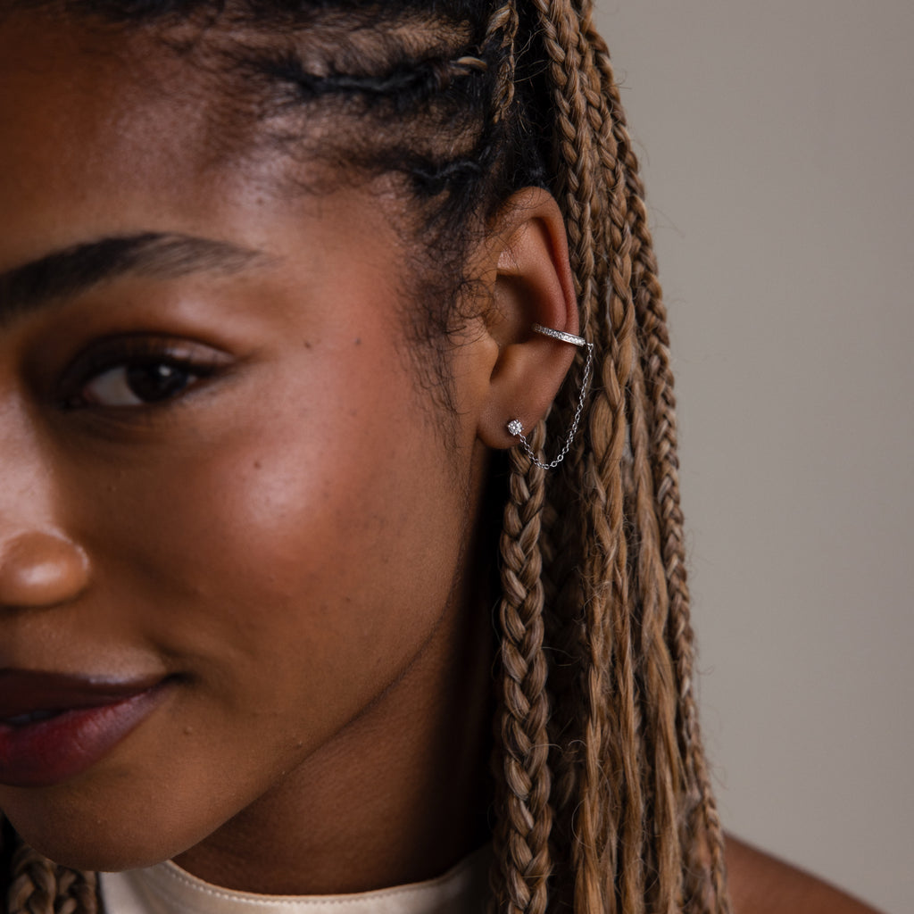 Close-up of a person with braided hair wearing a diamond studded earring with an ear cuff attached to a chain against a neutral background