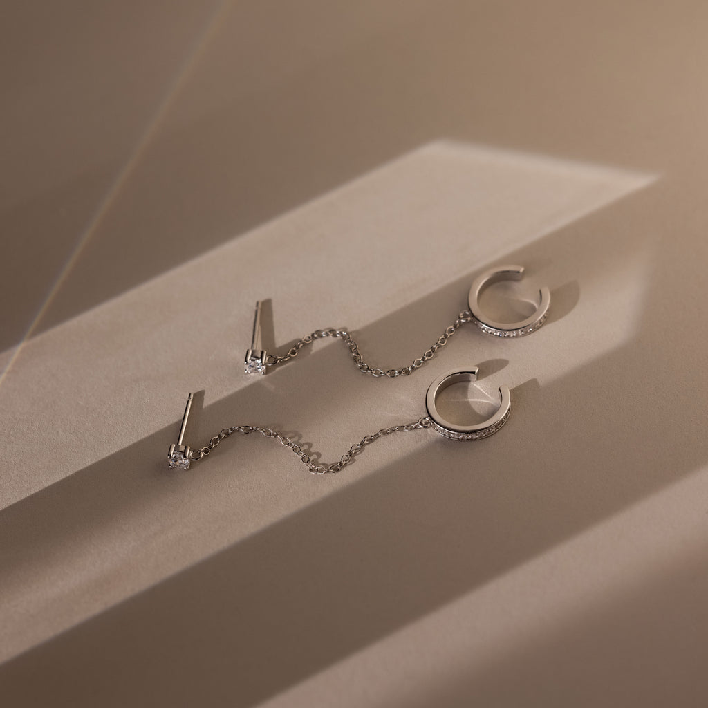 Silver diamond stud earrings with pave ear cuffs attached by a chain on a beige surface