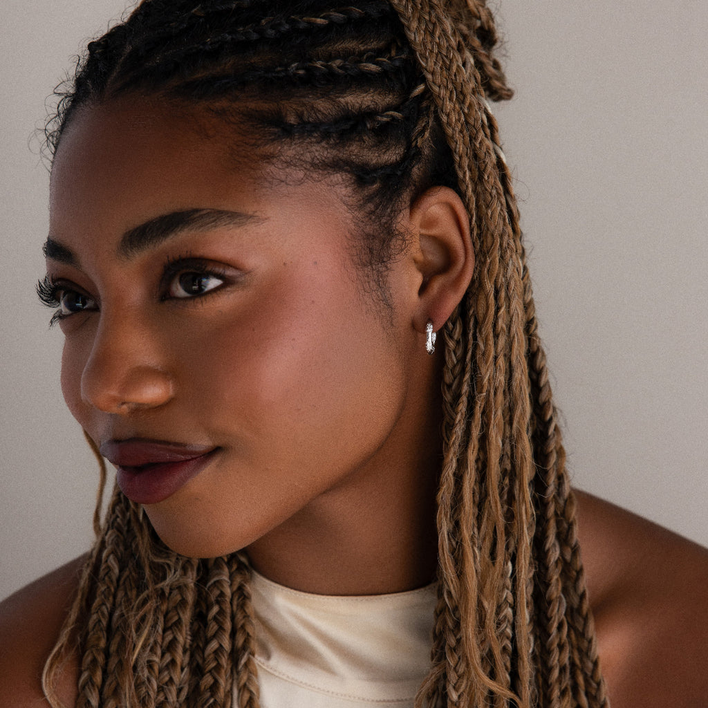 Woman with braided hair wearing silver diamond hoop earrings and a white top against a neutral background