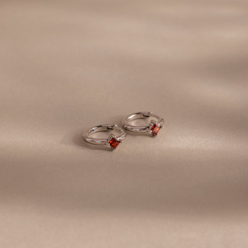 Two Athena Garnet Huggies—silver hoop earrings featuring small square red garnet gemstones—rest on a beige surface with soft shadows, making them perfect birthstone earrings and a thoughtful gift for loved ones.