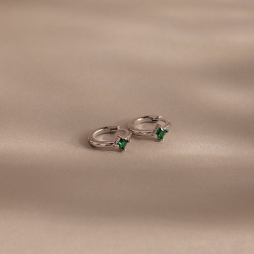 The Athena Emerald Huggies feature silver hoops with square green gemstones, displayed on a smooth beige surface—ideal for adding a stylish touch to any look or as personalized birthstone earrings.