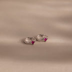 The Athena Ruby Huggies are silver hoop earrings featuring small square pink gemstones, adding a personalized jewelry touch as they rest elegantly on a light beige surface.