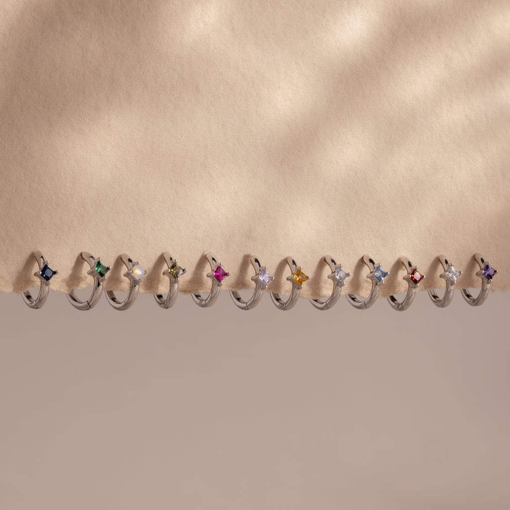 Twelve Athena Blue Zircon Huggies, silver rings with vivid blue gemstones, are displayed in a row on a beige background.