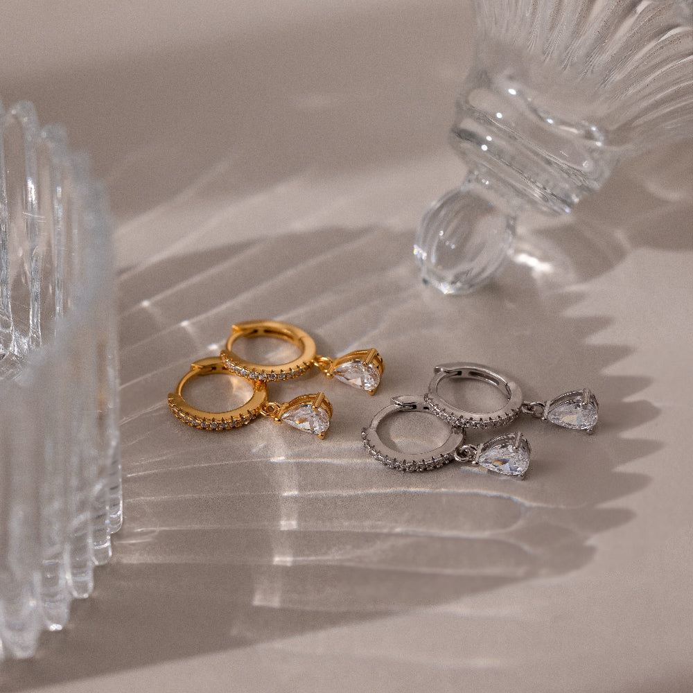 Two pairs of gold and silver hoop earrings with clear gemstones on a reflective surface.
