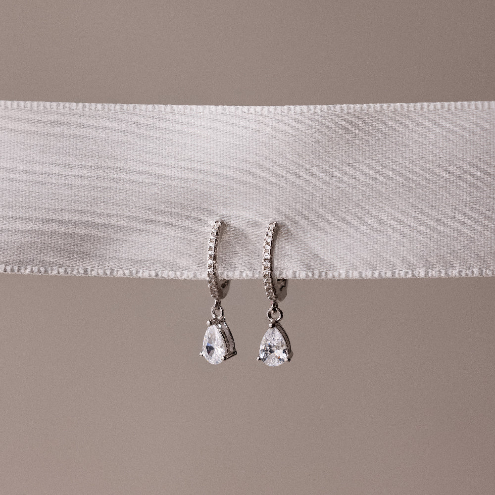 Pair of silver teardrop diamond hoop earrings on a white ribbon with a beige background