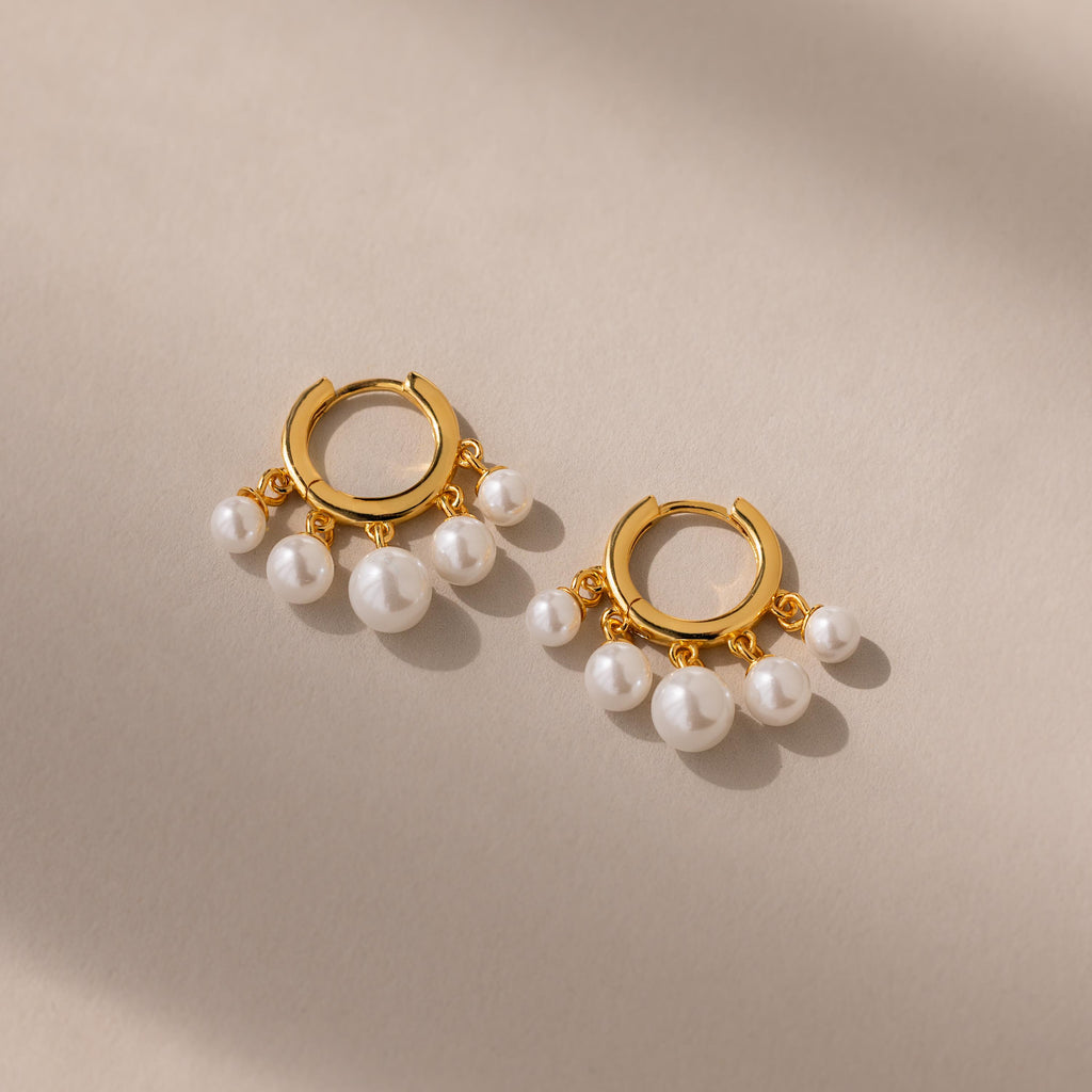 The Pearl Chandelier Huggies showcase gold hoops with dangling white pearl beads, elegantly displayed on a beige surface with soft shadows.