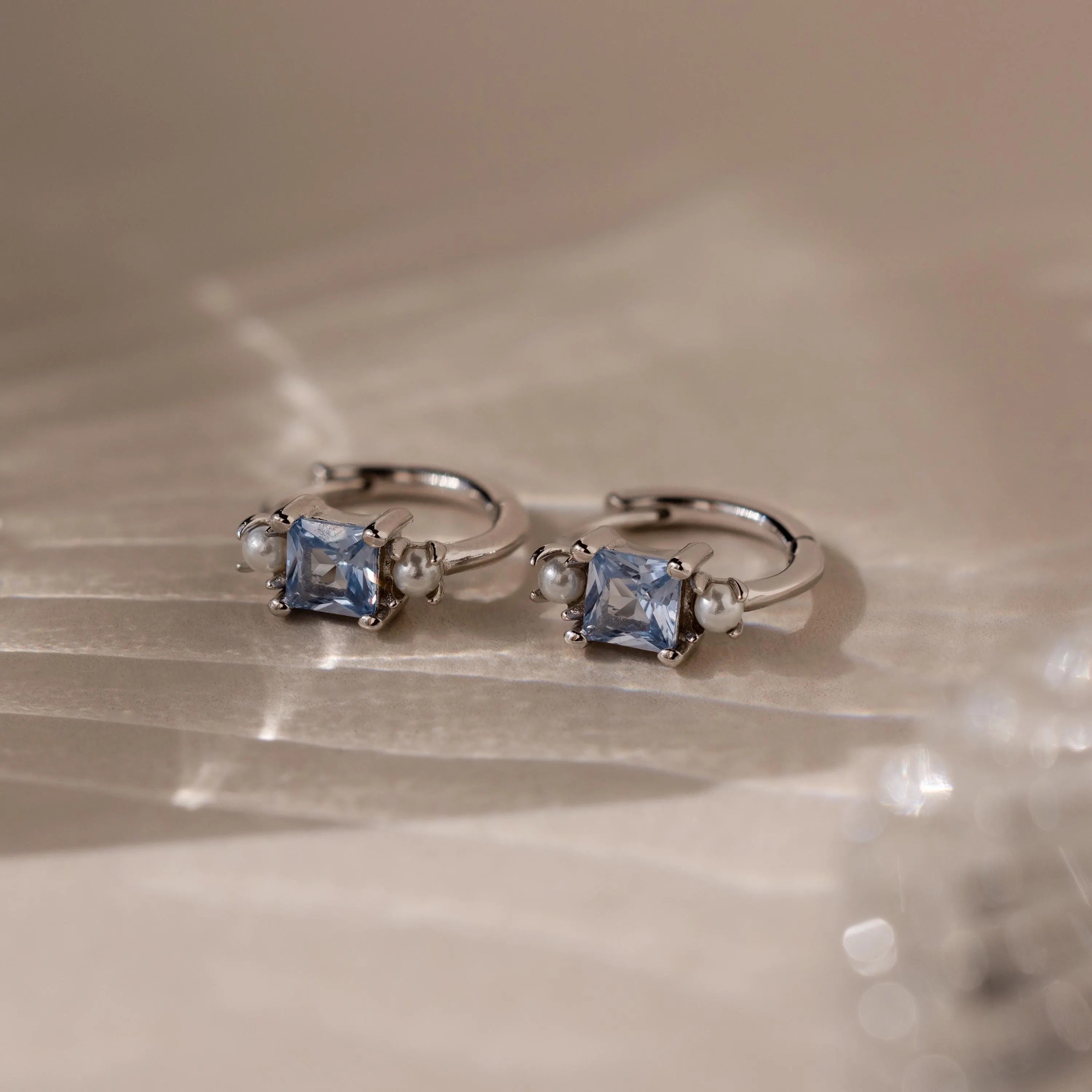 Aquamarine Pearl Huggies are silver hoop earrings featuring square blue gemstones and small pearls, making them stunning bridal jewelry or wedding earrings, beautifully displayed on a soft, light-colored surface.