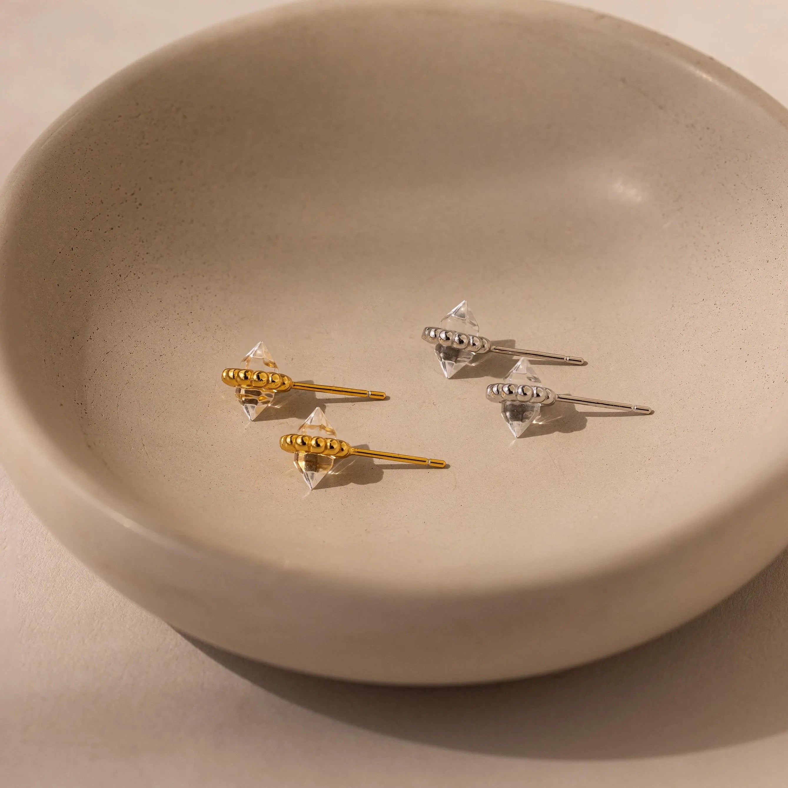 Two pairs of luminous earrings—one gold, one silver—are displayed in a round beige ceramic dish. The Beaded Herkimer Studs add subtle sparkle to the elegant arrangement.