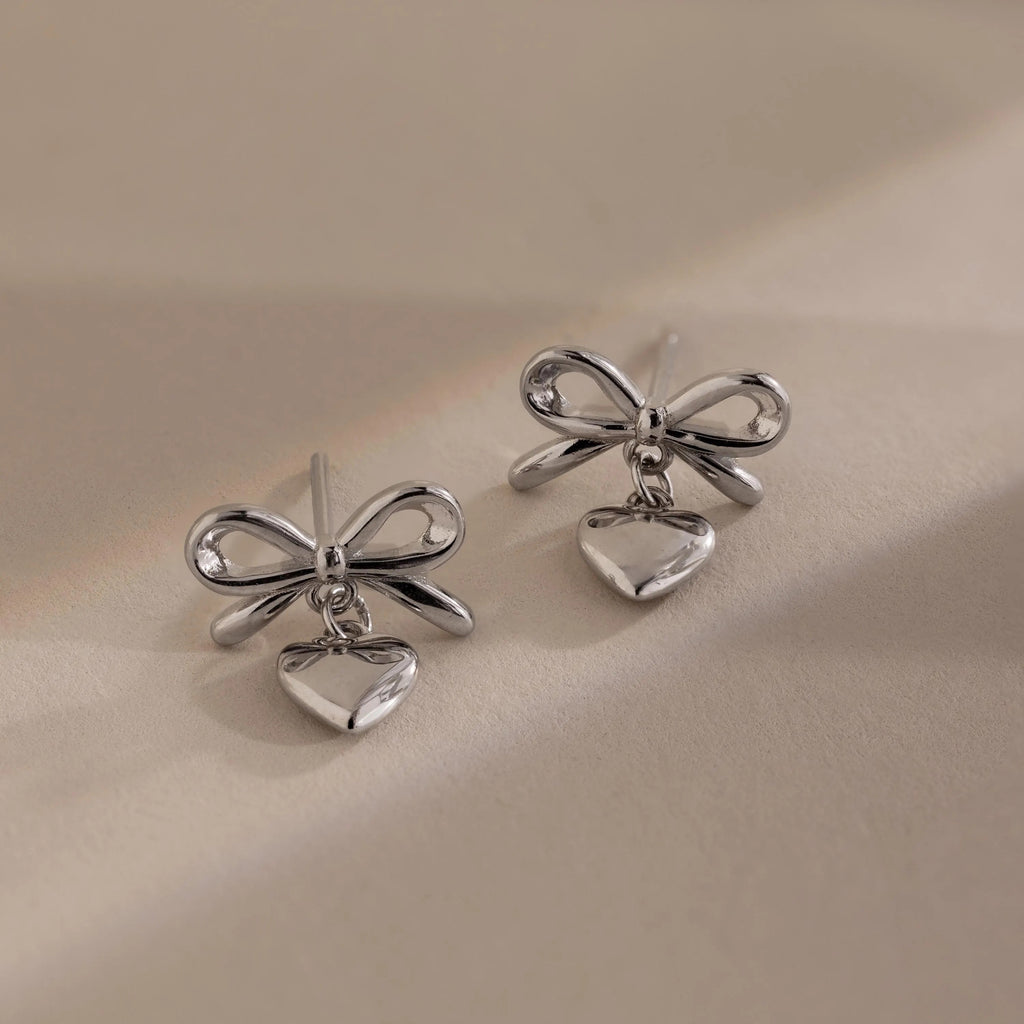 Two silver bow heart stud earrings with dangling heart charms placed side by side on a soft beige background.