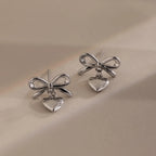 Two silver bow heart stud earrings with dangling heart charms placed side by side on a soft beige background.