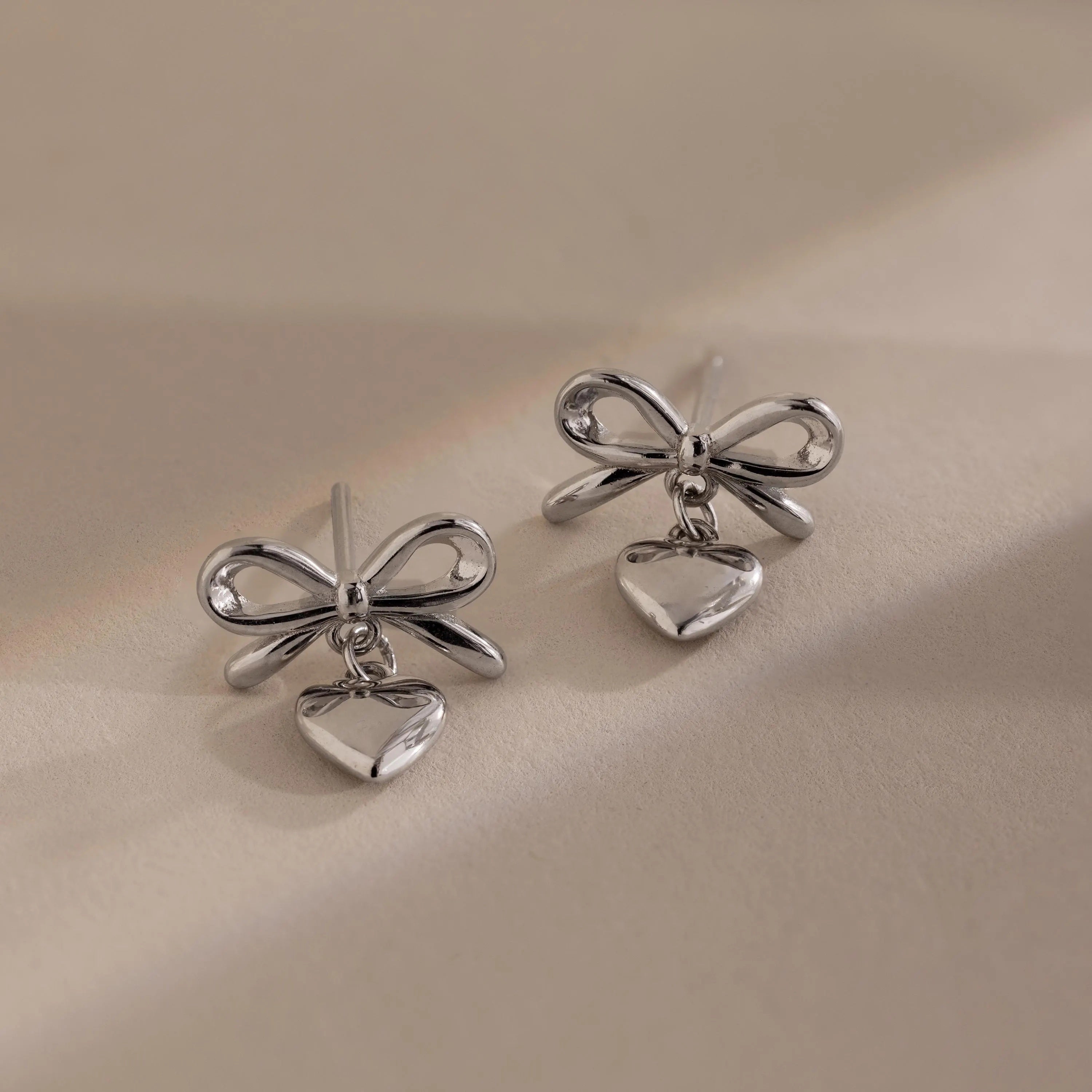 Two silver bow heart stud earrings with dangling heart charms placed side by side on a soft beige background.
