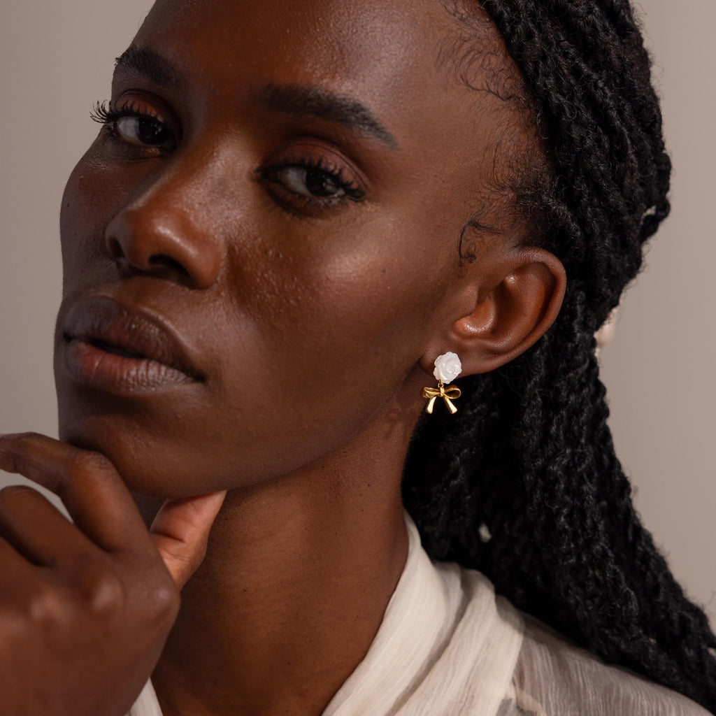 Woman gazing forward, wearing white rose and gold bow earrings, resting her chin on her hand.