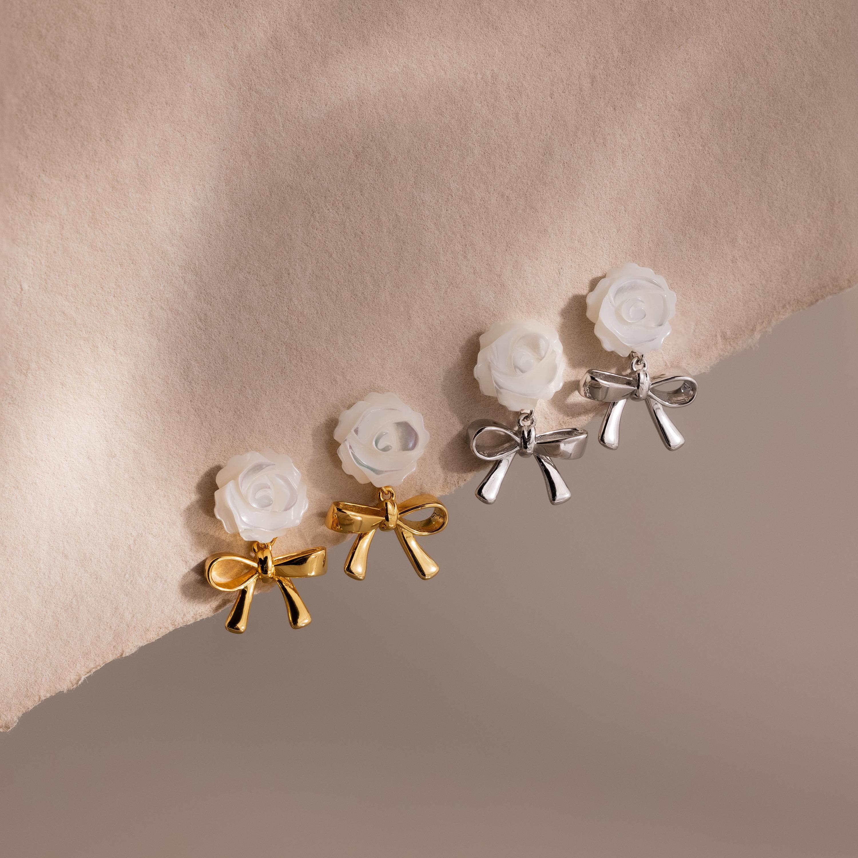 Two pairs of earrings—one gold, one silver—each with a white rose stud and metallic bow, arranged on soft taupe fabric.