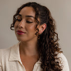 Woman with curly brown hair in a ponytail, wearing elegant Lace Diamond Huggies and a cream textured top, looking down.