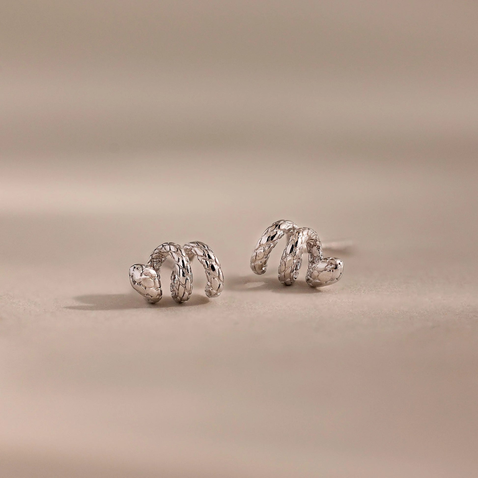 A pair of Twisted Snake Studs, silver and spiral-shaped, displayed on a soft beige surface with a blurred background—ideal for those who love elegant serpentine-inspired jewelry.