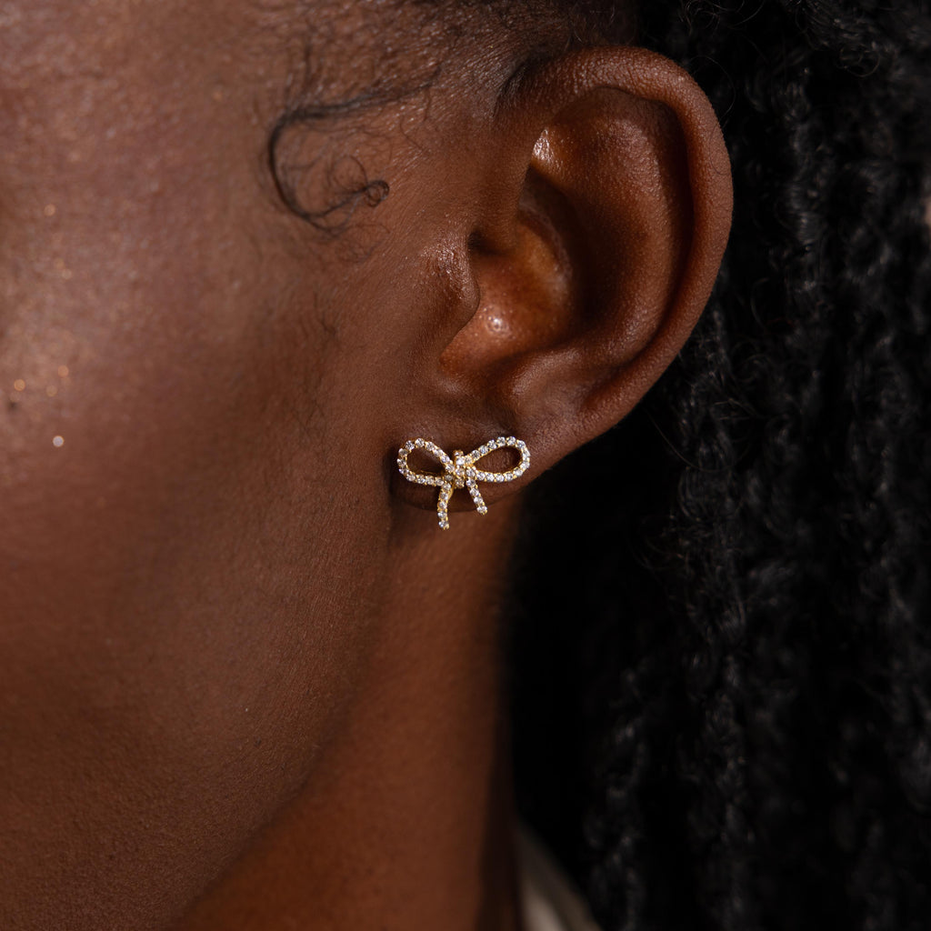 Close-up of model wearing a gold pavé bow stud earring, sparkling with tiny diamond stones and styled in a dainty ribbon shape.