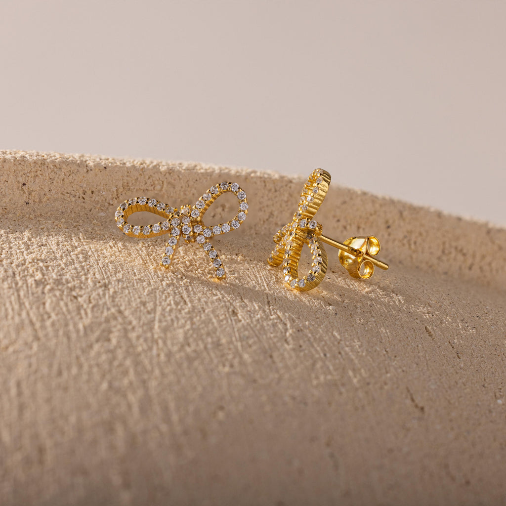 Close-up of gold pavé bow stud earrings resting on a textured beige surface, showcasing their sparkling ribbon design and detailed post backing.