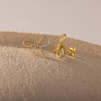 Close-up of gold pavé bow stud earrings resting on a textured beige surface, showcasing their sparkling ribbon design and detailed post backing.