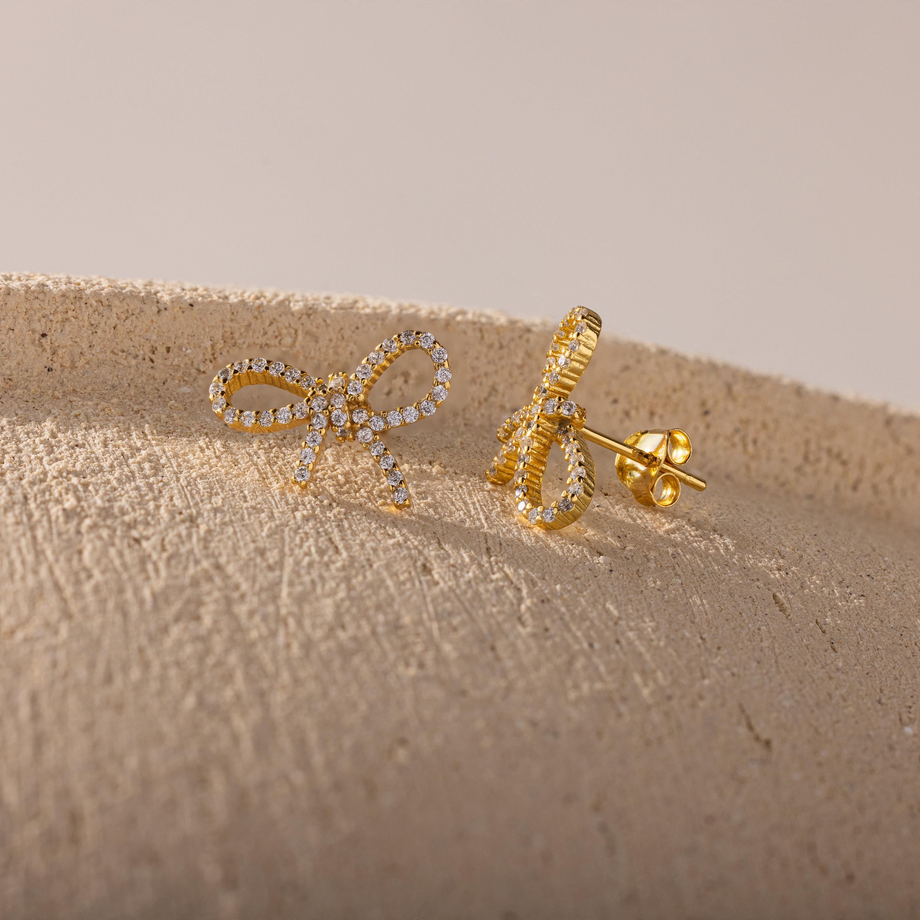 Close-up of gold pavé bow stud earrings resting on a textured beige surface, showcasing their sparkling ribbon design and detailed post backing.