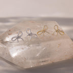Set of gold and silver pavé bow stud earrings displayed on a clear quartz crystal slab, showcasing their sparkling ribbon design under soft natural light.