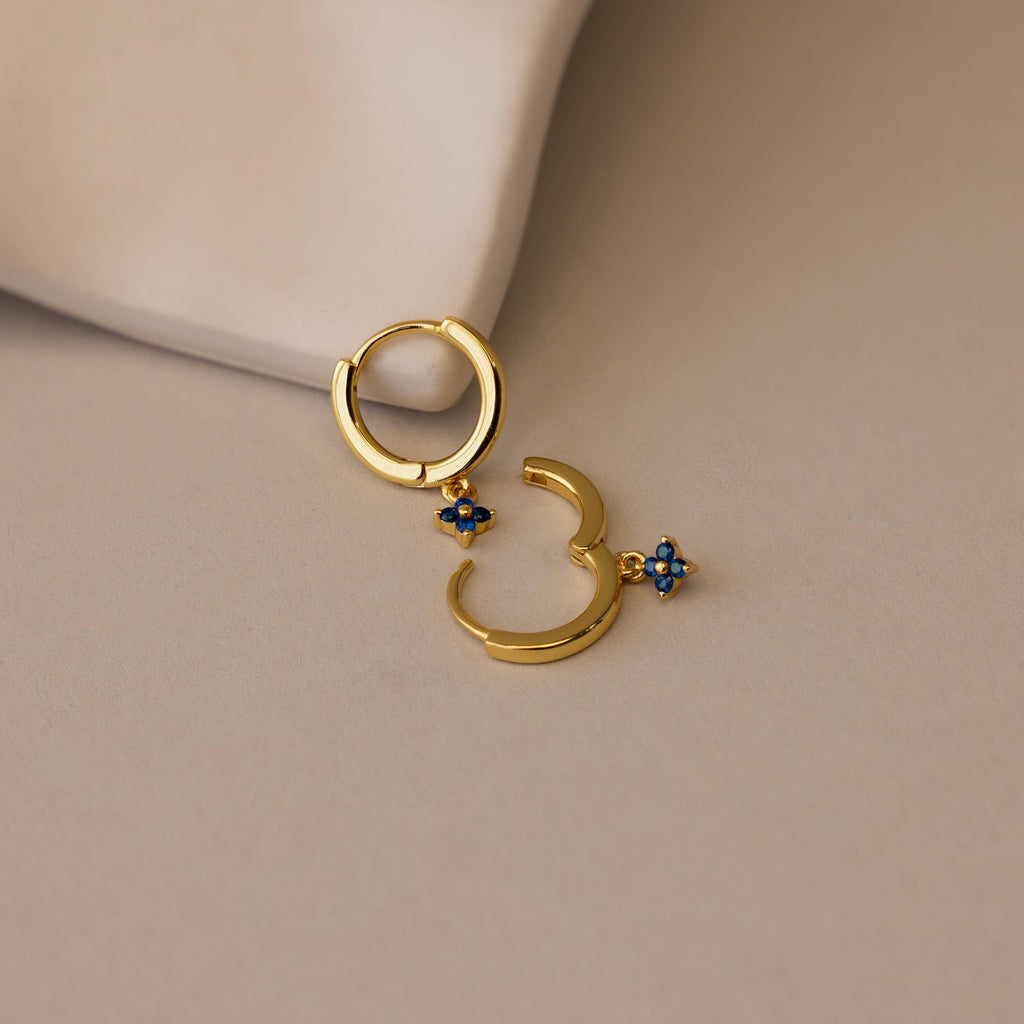 Ivy Birthstone Hoops—gold hoop earrings with small blue flower charms—add a delicate touch to any look, shown here on a light beige surface.