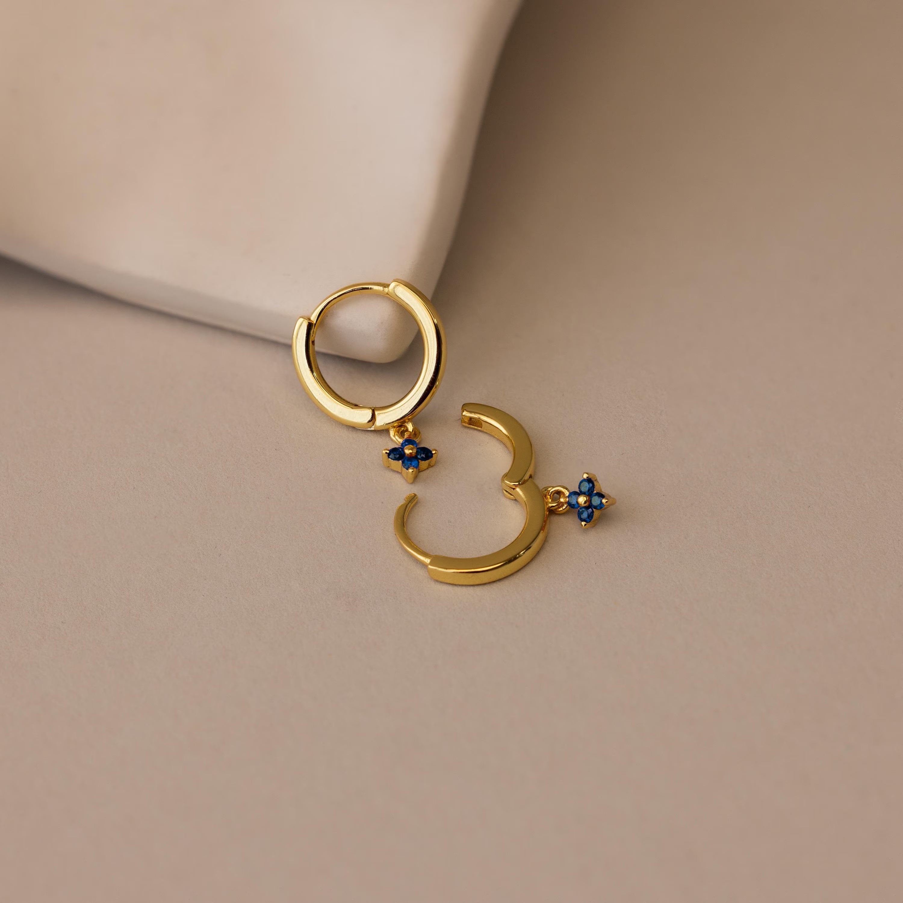 Ivy Birthstone Hoops—gold hoop earrings with small blue flower charms—add a delicate touch to any look, shown here on a light beige surface.