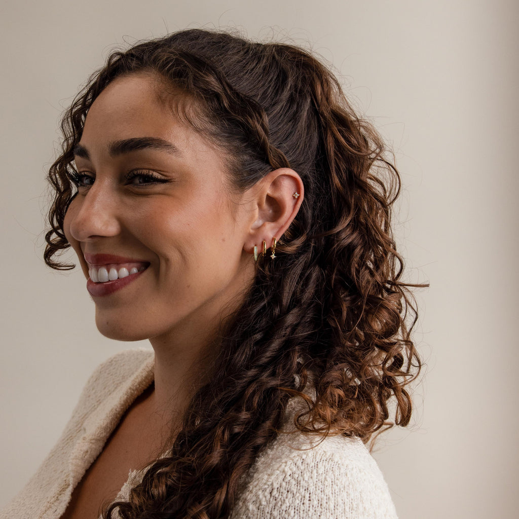 A smiling woman with curly hair and earrings, wearing a light sweater, faces left against a plain background while showcasing the elegant Ivy Birthstone Hoops.
