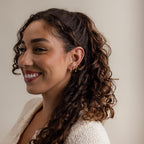 A smiling woman with curly hair and earrings, wearing a light sweater, faces left against a plain background while showcasing the elegant Ivy Birthstone Hoops.