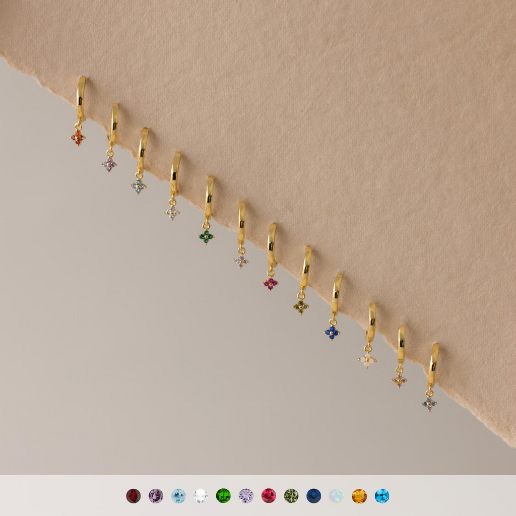 Twelve Ivy Birthstone Hoops with colorful gemstone charms are arranged diagonally on a beige and gray background.