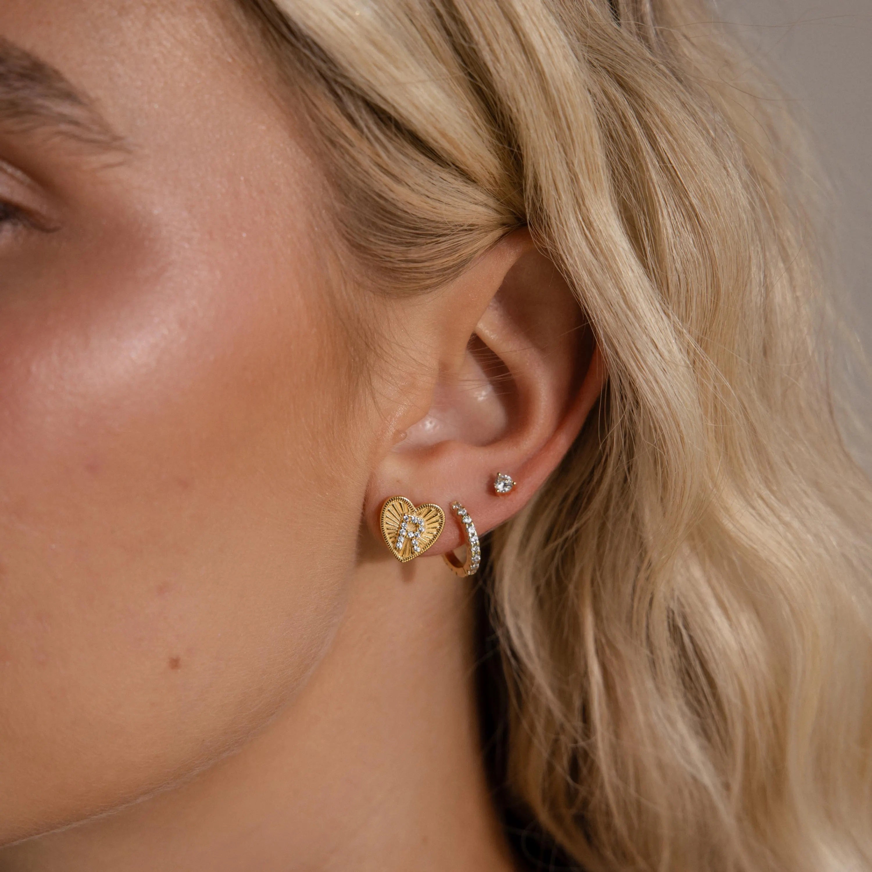Model wearing a gold Ribbed Heart Initial Earring with a pave letter R, stacked with a pave huggie hoop and dainty diamond stud.
