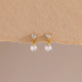 A close-up of a pair of 18k gold flat back earrings featuring heart-shaped studs with a dangling round crystal, displayed against a soft beige textured surface.