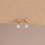 A close-up of a pair of 18k gold flat back earrings featuring heart-shaped studs with a dangling round crystal, displayed against a soft beige textured surface.