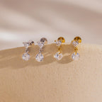 A side-by-side shot of two pairs each of the Heart Drop Flat Back Earrings in both 18k gold and sterling silver finishes, arranged on a light cream display ledge.