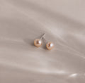 Pink Pearl Studs in a blush tone are showcased elegantly against a soft, light beige satin fabric background.
