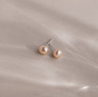 Pink Pearl Studs in a blush tone are showcased elegantly against a soft, light beige satin fabric background.