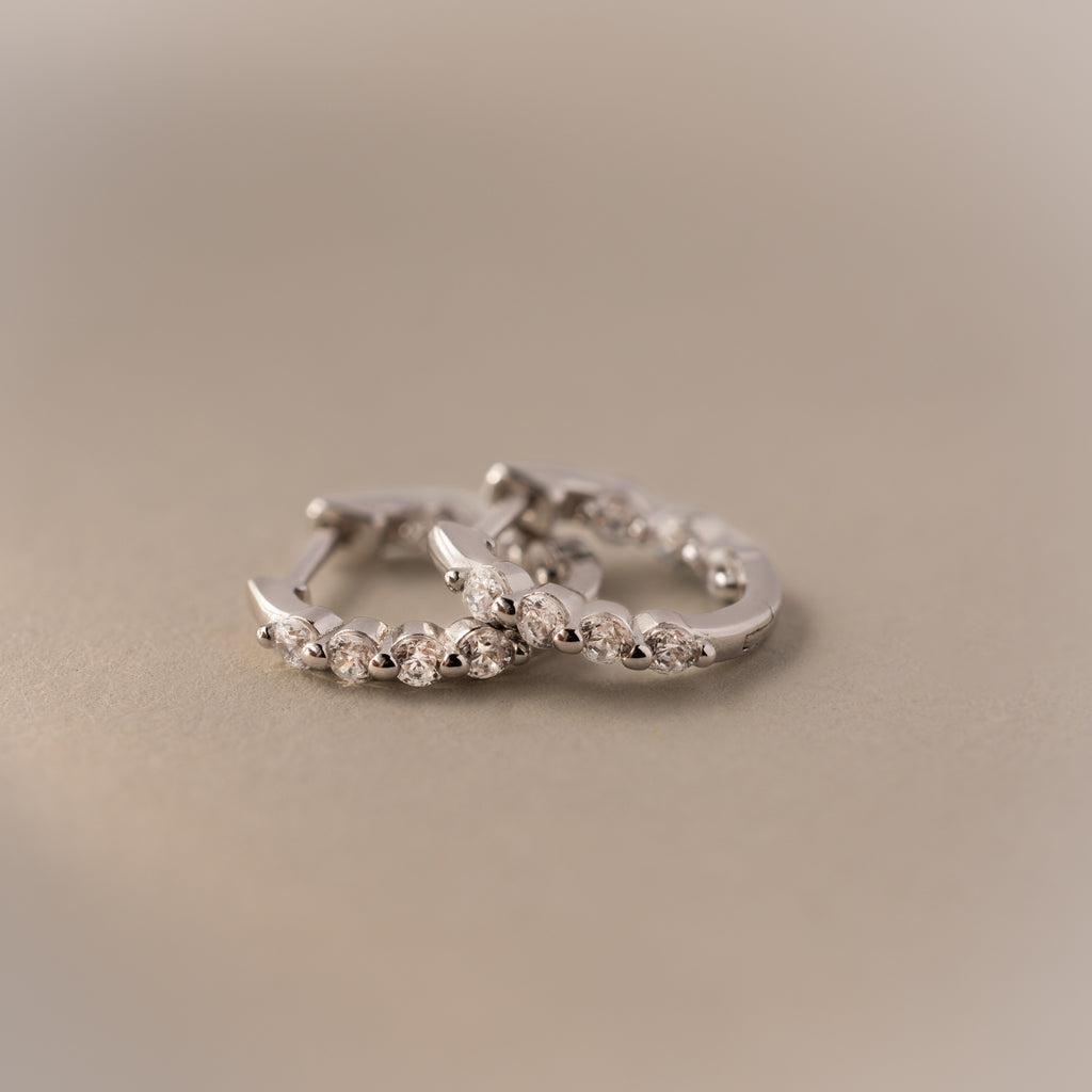Two Mini Birthstone Beaded Huggies—silver hoops with small embedded stones—are displayed on a soft beige surface.